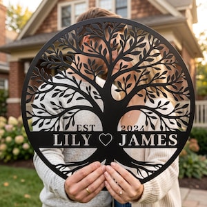 Op de afbeelding: Zwarte metalen wanddecoratie van een levensboom met de namen "LILY" en "JAMES", het jaar "2024" en een hartsymbool. Het boomontwerp is cirkelvormig met gedetailleerde bladeren en takken.