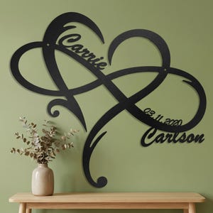 May include: A black metal wall art piece shaped like an infinity symbol intertwined with a heart. The design features the name Carrie, the date 03.11.2020, and the name Carlson integrated into the flowing, continuous lines of the metal structure.