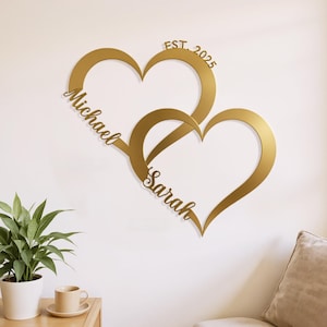May include: A gold-colored wall hanging featuring two intertwined heart shapes. The names "Michael" and "Sarah" are inscribed on the hearts, with "EST. 2025" above. The design is mounted on a white wall, suggesting a decorative piece for a home.