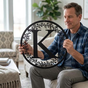 May include: A black metal monogram letter "K" in a circular frame with floral accents. The decorative wall art is held by a person in a blue plaid shirt. The metal sign is designed for indoor or outdoor use.