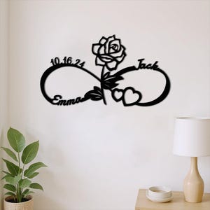 May include: Black metal wall art depicting an infinity symbol with a rose, two hearts, and the names "Emma" and "Jack." The date "10.16.24" is also included. Ideal for home decoration.