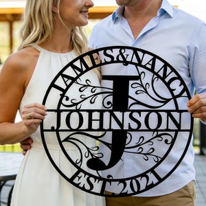 May include: Black metal family name sign with the names "JAMES & NANCY" and "JOHNSON" with the letter "J" in the center. The sign also includes "EST 2021" and decorative floral elements.