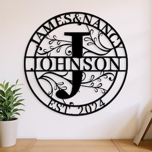 May include: Black metal family name sign featuring the names "JAMES & NANCY" and "JOHNSON" in a circular design. The sign includes the year "EST 2024" and decorative floral elements.