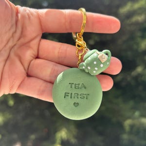 May include: A green keychain with a gold ring. The keychain features a round charm with the words "TEA FIRST" and a small heart, plus a miniature teapot with white polka dots. The background is blurred.