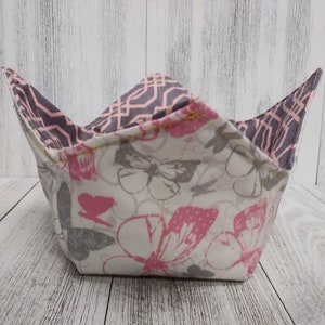 Può includere: Una ciotola quadrata in tessuto con un motivo a farfalle rosa e grigio su sfondo bianco. La fodera interna presenta un design geometrico grigio con accenti rosa. La ciotola è progettata per contenere una ciotola di zuppa calda o altro cibo.