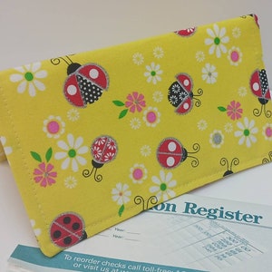 May include: Yellow checkbook cover with a ladybug and flower pattern. The cover is made of fabric and has a stitched edge.