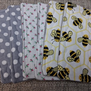 Puede incluir: Cuatro tiras de tela con diferentes patrones. Una tira tiene un fondo gris con lunares blancos. Otra tira tiene un fondo blanco con pequeñas flores rosas. La tercera tira tiene un fondo blanco con un patrón de panal y abejas amarillas y negras. La cuarta tira tiene un fondo blanco con un patrón de panal y abejas amarillas y negras.