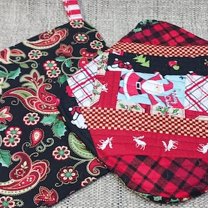 Op de afbeelding: Twee kerst-thema pottenlapjes. Eén is zwart met een paisley-patroon en de andere is rood en zwart met een patchwork-ontwerp met een kerstmanfiguur.