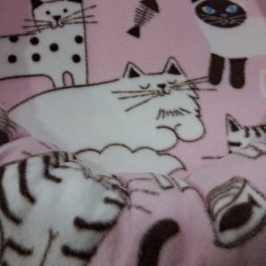 May include: Pink cat bed with a repeating pattern of cartoon cats, fish bones, and cat food. The bed has a soft, plush texture and a quilted design. The cats are in various poses, with white and black markings.
