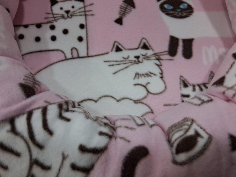 May include: Pink cat bed with a repeating pattern of cartoon cats, fish bones, and cat food. The bed has a soft, plush texture and a quilted design. The cats are in various poses, with white and black markings.