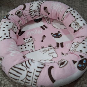 May include: Pink and white cat bed with a circular design. The bed features a soft, plush material with a repeating pattern of cartoon cats, fish bones, and the word "MEOW". The bed has a raised, padded rim.
