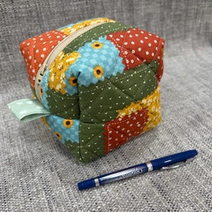 May include: A quilted fabric cube pouch featuring a patchwork design of orange, yellow, green, and light blue floral patterns. The pouch has a beige zipper closure and a light green polka dot fabric tab. A blue ballpoint pen is placed next to it.