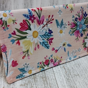 May include: A pink fabric wallet with a floral pattern featuring white, yellow, pink, blue, and green flowers. The wallet has a flap closure and is made of a soft, textured fabric.