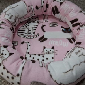 May include: Pink cat bed with a plush, rounded design. The bed features a repeating pattern of various cat illustrations in black and white, with the word "MEOW" printed in white. The bed is designed for pets.