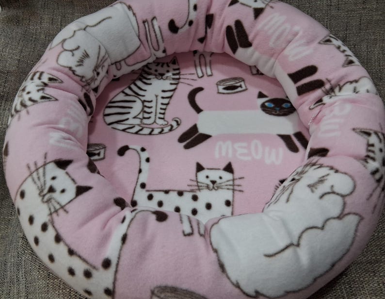 May include: Pink cat bed with a plush, rounded design. The bed features a repeating pattern of various cat illustrations in black and white, with the word "MEOW" printed in white. The bed is designed for pets.