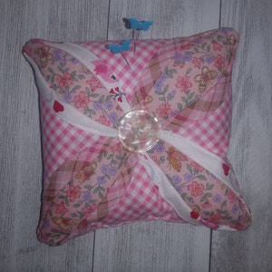 May include: A pink and white pincushion with a floral print and gingham pattern. It has a pearl button in the center and is decorated with butterflies and hearts.