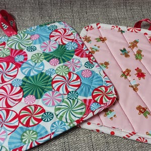May include: Two quilted pot holders. One is pink with a pattern of gingerbread men. The other is white with a pattern of red and green peppermint swirls.