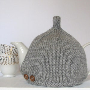 May include: Grey knitted teapot cosy with brown buttons. The cosy is on top of a white teapot with a white handle and spout. There are three white teacups stacked next to the teapot.