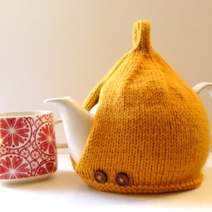 May include: A yellow knitted teapot cozy with two brown buttons. The cozy is on a white teapot with a white mug next to it.