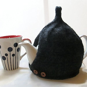 May include: A black knitted teapot cozy with two brown buttons. The cozy is on a white teapot with a white mug with black and red accents next to it.
