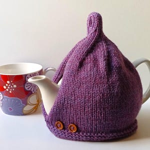 May include: A white ceramic teapot with a purple knitted tea cozy and two brown buttons. A floral patterned mug is next to the teapot. The cozy is a deep purple color.