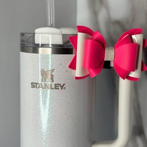 Könnte beinhalten: Ein weißer Glitzer-Stanley-Becher mit Henkel und einem durchsichtigen Strohhalm. Eine leuchtend rosa-weiße Schleife ist am Henkel befestigt. Das Stanley-Logo ist auf der Vorderseite des Bechers zu sehen.