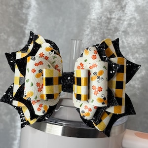 May include: A decorative hair bow with a black and yellow checked pattern, and a central section decorated with bees and floral motifs. The bow has black glitter detailing and is placed on a white tumbler.