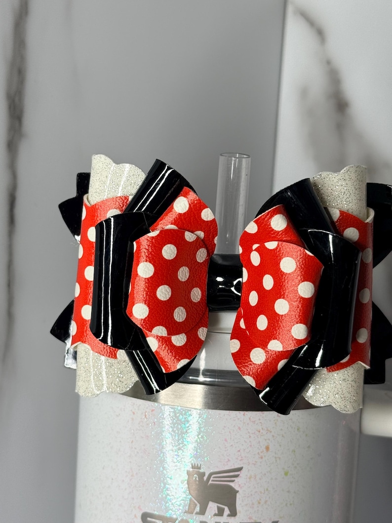 May include: A red and white polka dot bow with black detailing sits atop a white tumbler. The bow is made of a shiny material and has a glittery white base. A clear straw is visible.
