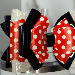 May include: Close-up of a decorative bow featuring red material with white polka dots, black accents, and a glittery white section. The bow is attached to a white tumbler, creating a playful and festive accessory.
