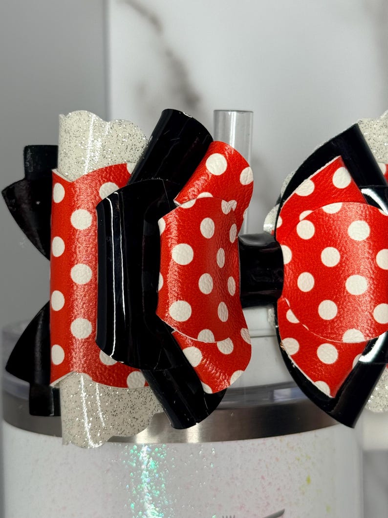 May include: Close-up of a decorative bow featuring red material with white polka dots, black accents, and a glittery white section. The bow is attached to a white tumbler, creating a playful and festive accessory.