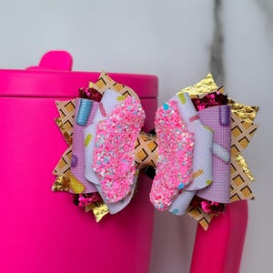 May include: A decorative hair bow featuring a layered design with a waffle cone pattern, pink glitter accents, and small colorful sprinkles. The bow is constructed from multiple textured materials including gold metallic, dark red glitter, and light purple fabric.