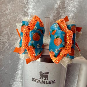 May include: A decorative hair bow featuring a pumpkin design. The bow has layers of orange glitter, blue fabric with pumpkin prints, and white accents. It is attached to a white Stanley mug.