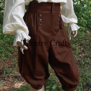 May include: Brown, high-waisted breeches with decorative buttons, part of a costume. The breeches have a gathered, voluminous shape and are paired with a cream-coloured blouse with ruffled sleeves. The text "Theatre Crafts" is visible on the breeches.