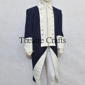 May include: A navy blue long coat featuring contrasting cream-colored lapels, cuffs, and front panels. The garment includes two vertical rows of silver-toned buttons along the front opening. The text Theatre Crafts is centered on the image.