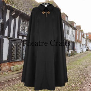 May include: A black cape with a collar and decorative gold-colored clasps. The cape is long and flowing, with the text "Theatre Crafts" visible on the front. The background features a cobblestone street and historic buildings.