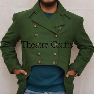 Op de afbeelding: Groene wollen jas met een dubbele rij knopen en goudkleurige knopen. De jas heeft een unieke snit en draagt de tekst "Theatre Crafts". Het model draagt er een tealkleurig shirt onder.