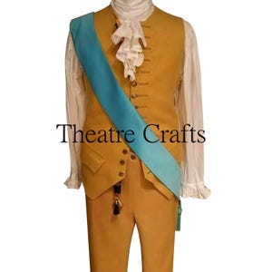 May include: A mustard yellow waistcoat and matching trousers paired with a white long-sleeved shirt featuring a ruffled jabot. A bright turquoise sash is worn diagonally across the chest. The text Theatre Crafts is centered on the image.