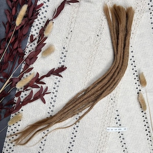 Op de afbeelding: Een bundel lichtbruine dreadlocks, diagonaal geplaatst op een wit, patroonrijk oppervlak. De dreadlocks zijn getextureerd en lijken van synthetisch haar te zijn. Decoratieve gedroogde planten met bordeauxrode bladeren en beige accenten zijn op de achtergrond.