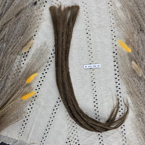 Op de afbeelding: Een set bruine synthetische dreadlocks met een getextureerd, gematteerd uiterlijk. De dreadlocks zijn aan de bovenkant samengebundeld en lopen taps toe naar de uiteinden. Een klein wit rechthoekig label met de zwarte tekst 60 10-11 0126 137 is zichtbaar.