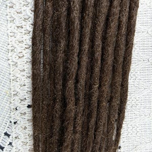 Puede incluir: Primer plano de un grupo de rastas de color marrón oscuro. Las rastas están muy juntas, mostrando un aspecto texturizado y natural. El fondo es una tela blanca estampada.