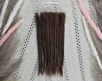 May include: A set of brown dreadlocks arranged in a vertical row, featuring a small white rectangular label with the text 25 8-9 0126 118 positioned above the hair strands. The dreadlocks have a textured, matted appearance and a uniform dark brown colour.