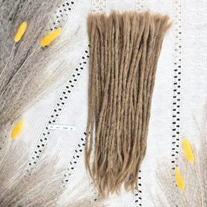 Op de afbeelding: Een bundel lichtbruine synthetische dreadlocks met losse, golvende uiteinden. Een klein wit rechthoekig label is zichtbaar bij de bovenkant, met de gedrukte tekst 35 6-7 0126 28. De dreadlocks zijn verticaal in een uniforme formatie gerangschikt.