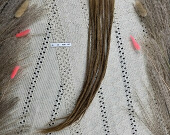 May include: A collection of brown dreadlocks, displayed on a cream-coloured, patterned surface. The dreadlocks are a light brown shade and look to be synthetic. The background features dried plants with pink and beige accents.