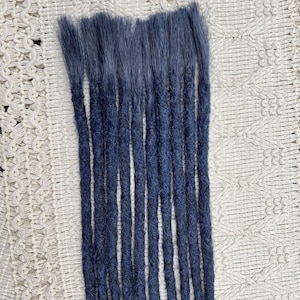 May include: A collection of dark blue dreadlocks, displayed vertically against a textured, off-white backdrop. The dreadlocks have a faded appearance at the ends, creating a subtle ombre effect. The overall aesthetic suggests a bohemian or alternative style.