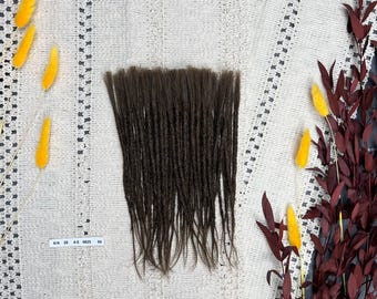 May include: A collection of dark brown dreadlocks, displayed on a white, textured surface. The dreadlocks are long and thin, with a natural, textured appearance. Yellow and burgundy decorative elements are arranged around the dreadlocks. A small tag with text is visible.