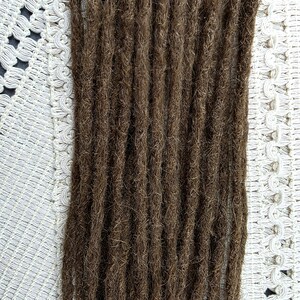 May include: Close-up of brown dreadlocks, tightly twisted and textured. The dreadlocks are arranged vertically, showcasing their individual strands and overall thickness. The background is a white, patterned fabric.