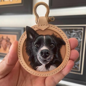 Pode incluir: Um porta-chaves redondo de couro bege com um retrato detalhado de um cão preto e branco. O cão tem uma expressão focada, com uma mancha branca no nariz. O porta-chaves tem um fecho cor de latão.