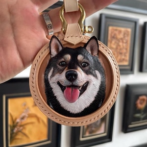 Pode incluir: Um porta-chaves redondo de couro castanho com um retrato de cão Shiba Inu sorrindo. O cão tem pelo preto e branco com uma língua rosa. O porta-chaves tem um fecho dourado e uma correia de couro castanho.