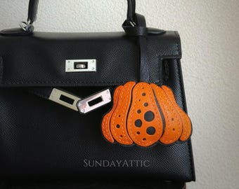 May include: Black leather handbag with a top handle and silver hardware. An orange pumpkin-shaped leather charm with black spots hangs from the handle. The text "SUNDAYATTIC" is visible on the bag.