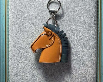 May include: A leather horse head keychain with a silver keyring. The horse head is tan with blue fringe and white stitching. The keychain is displayed in a blue framed shadow box with a gray background.